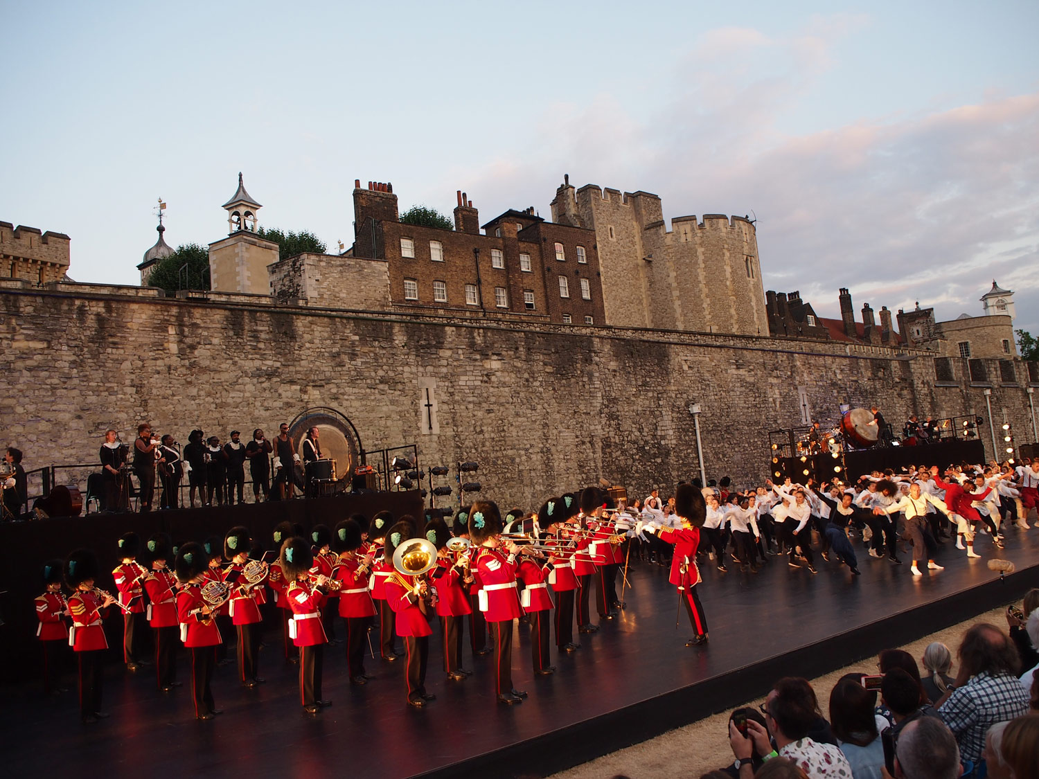 In front of a large castle wall is a black stage. Raised on a platform are a band, all dressed in black. On the stage, a military brass marching band are dress in red with black bearskin hats. Also on the stage are dozens of young people, dancing.