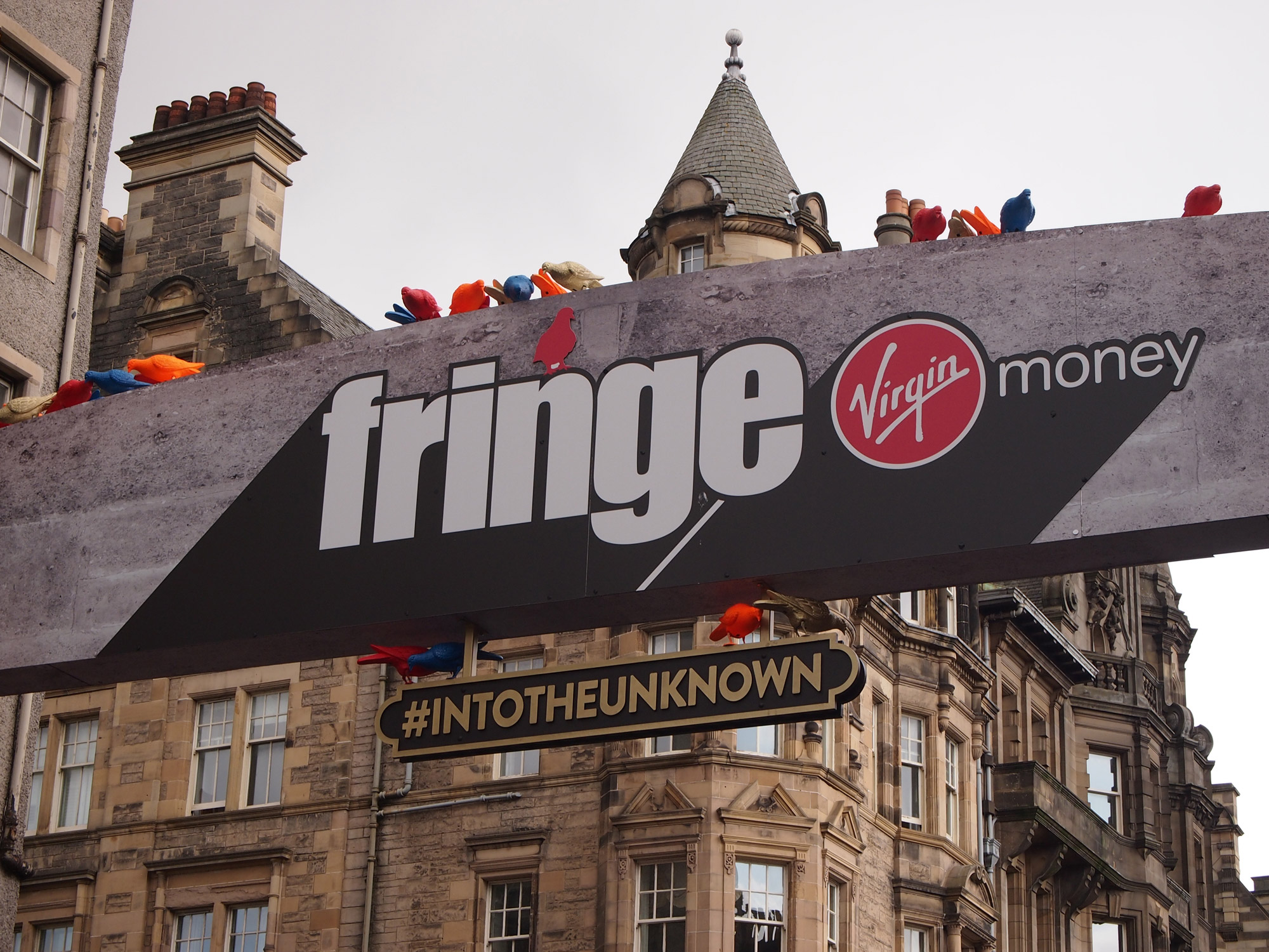 A sign hangs across a road, it is covered in multi-coloured plastic pigeons. The sign reads 'Fringe, into the unknown'. Behind it we see gothic buildings.