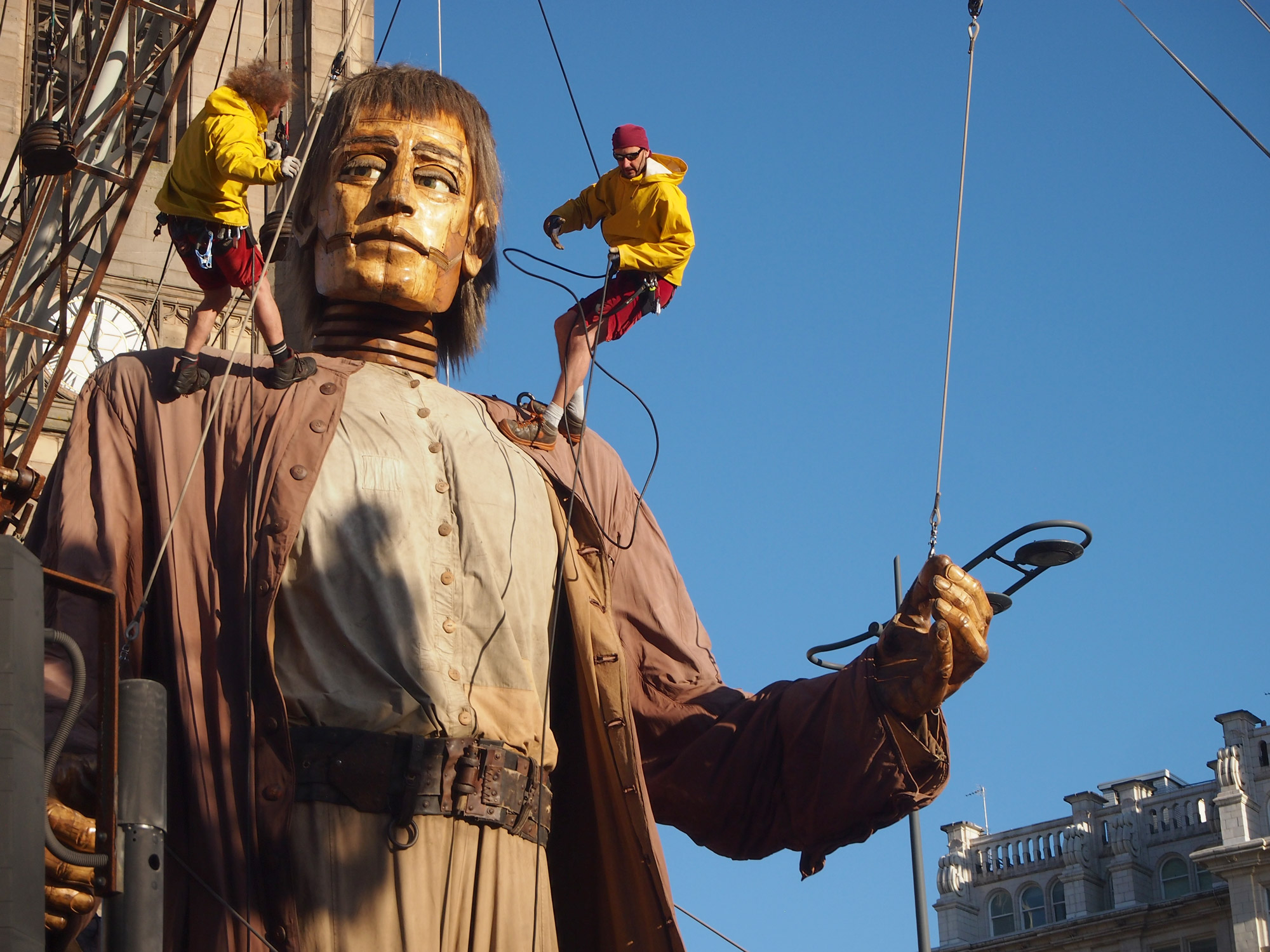 On a giant mannequin, two men hang on ropes. The giant appears to be looking at one of the men.