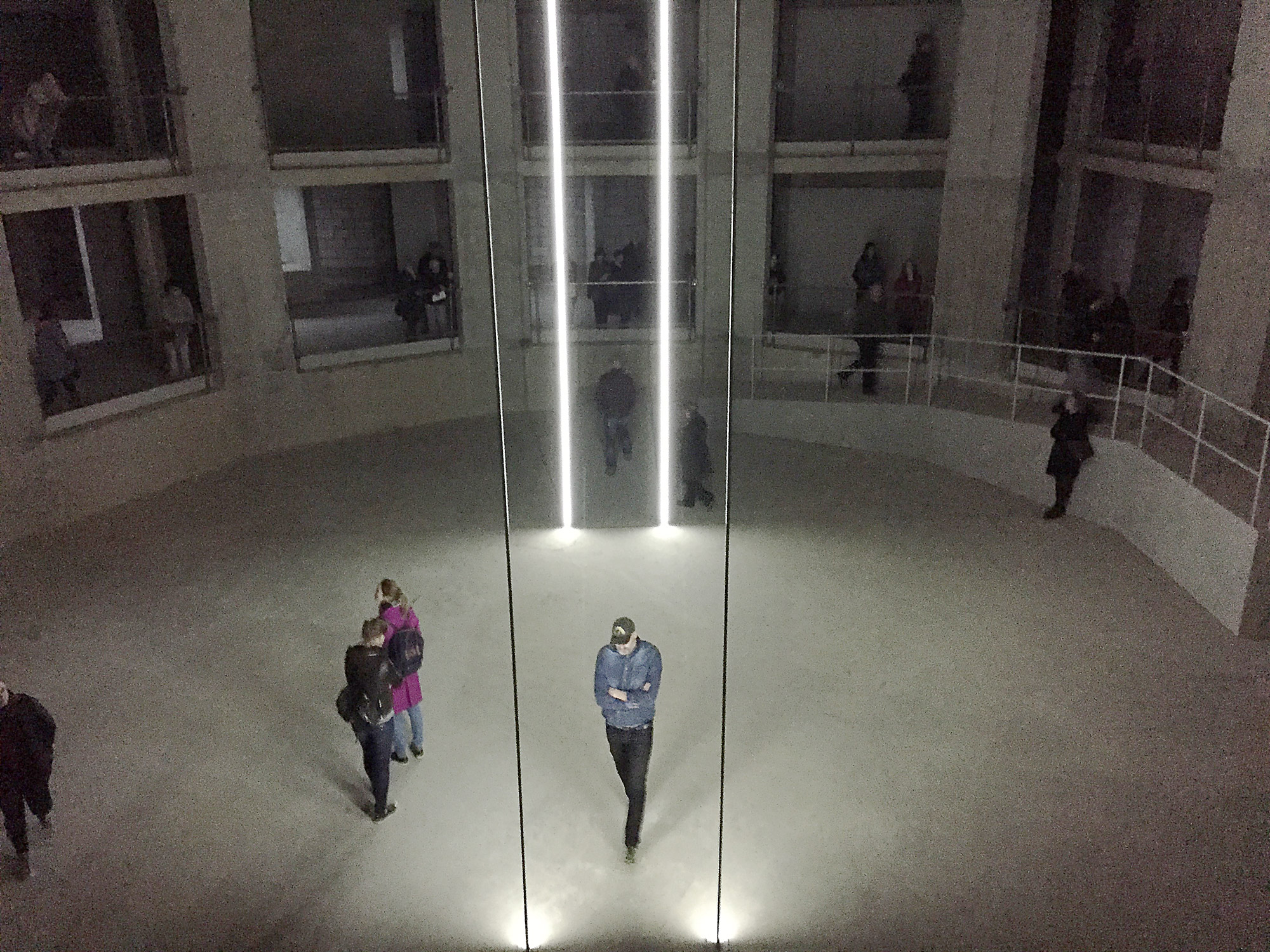 People walk around a large concrete space with apertures on two floors. There are vertical beams of light, cast up from the floor.
