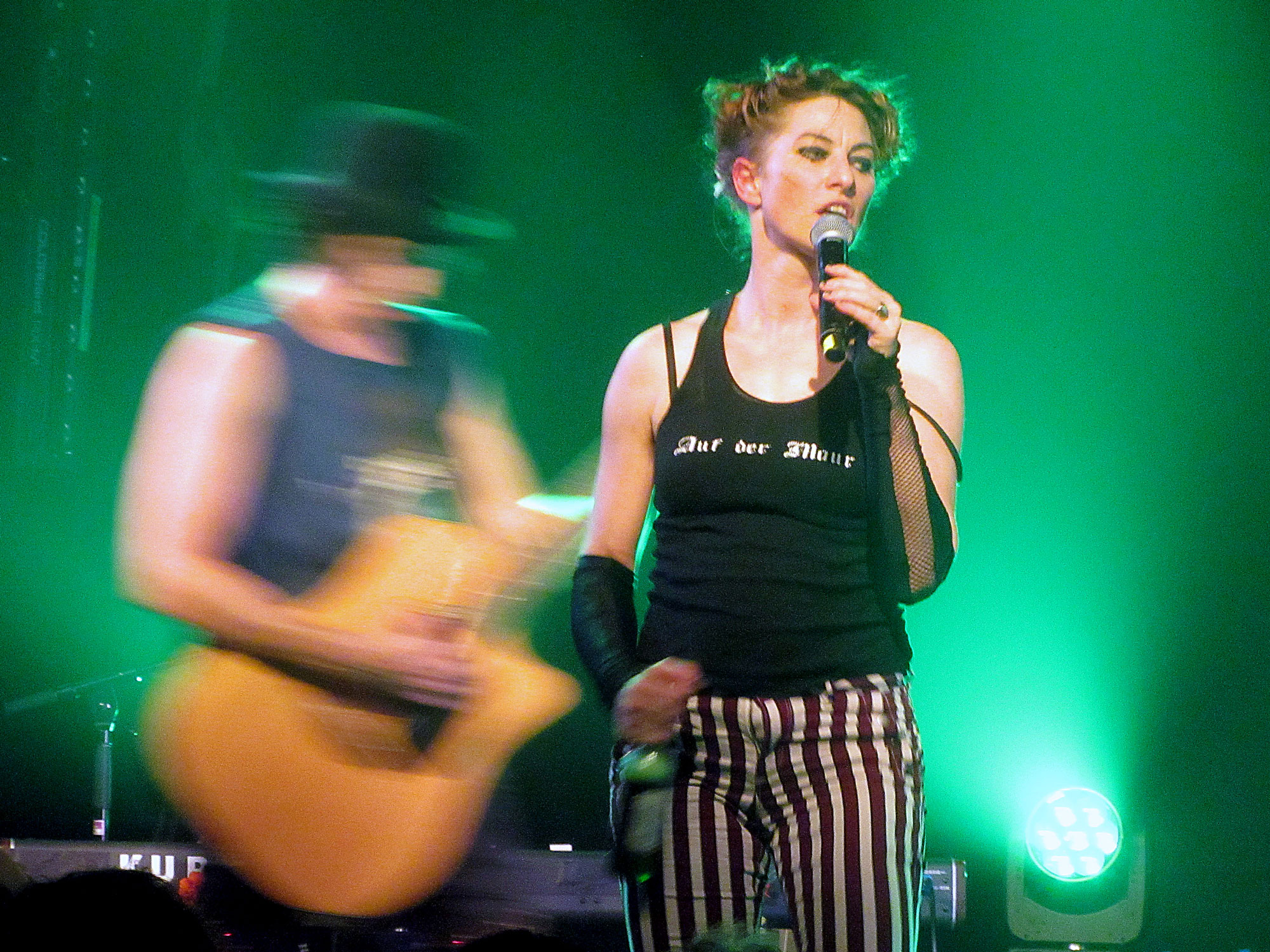 On a green-lit stage, stands a woman dressed in a black vest and stripy trousers. In one hand she holds a microphone, in the other a bottle of beer. Behind her, the blurred figure of a man playing acoustic guitar. He wears a bowler hat.