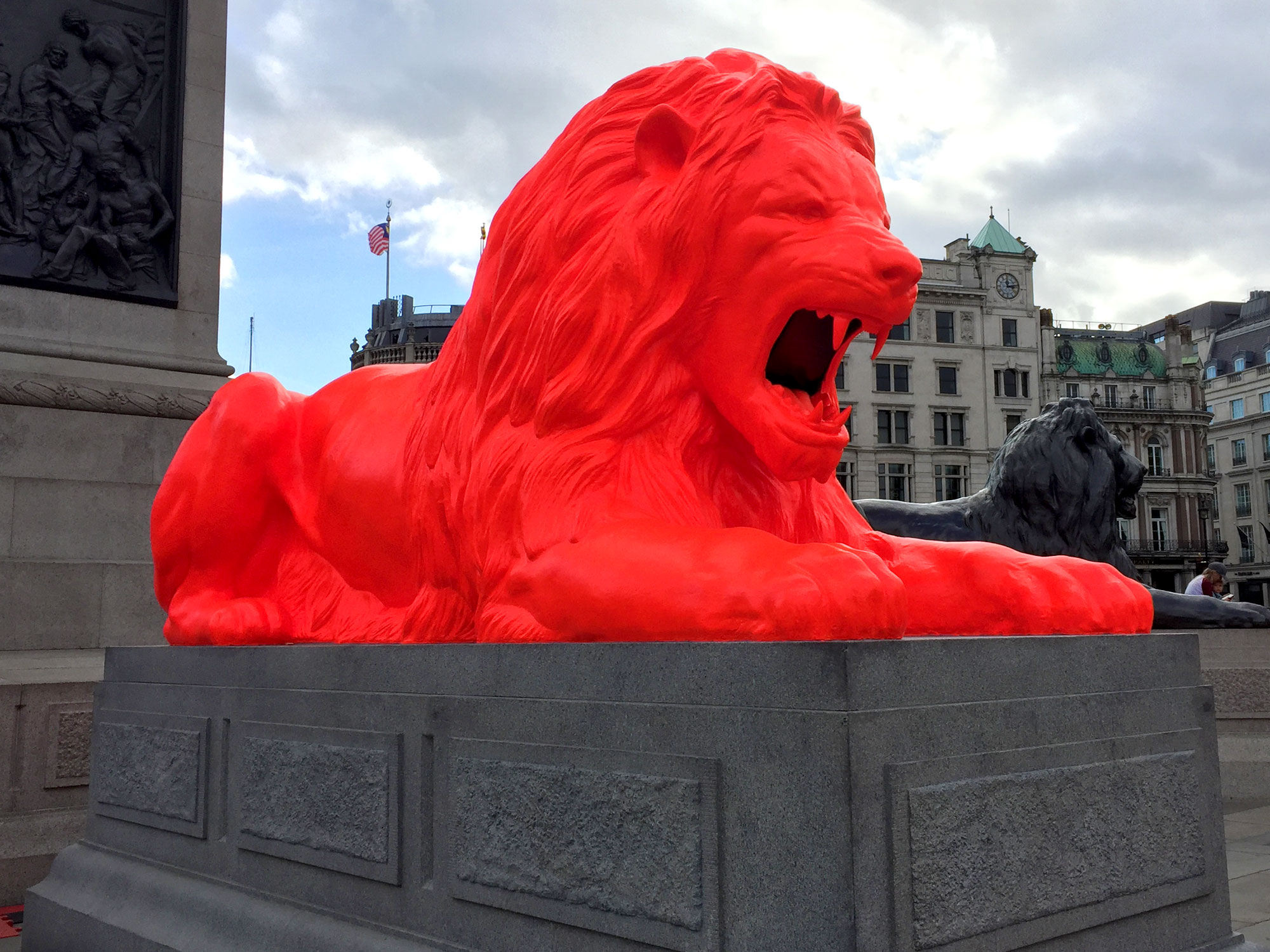 On a grey stone plinth sits a reclining line sculpture, painted in fluorescent red paint. Behind it a similar black lion and behind that is a London skyline.