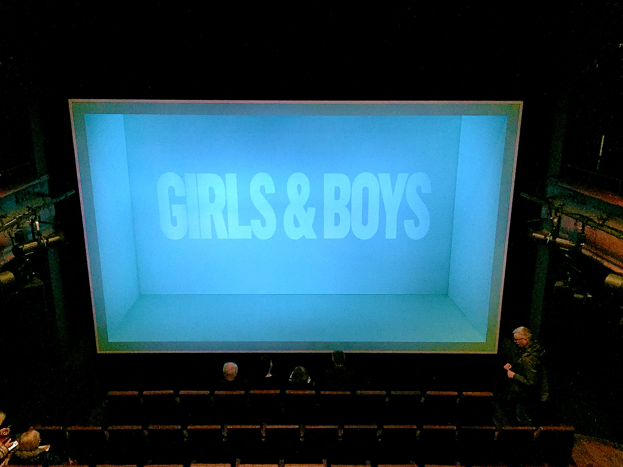 In front of rows of theatre seats, a green-lit box is open on one side towards us. At the rear of the box are the words Girls & Boys.
