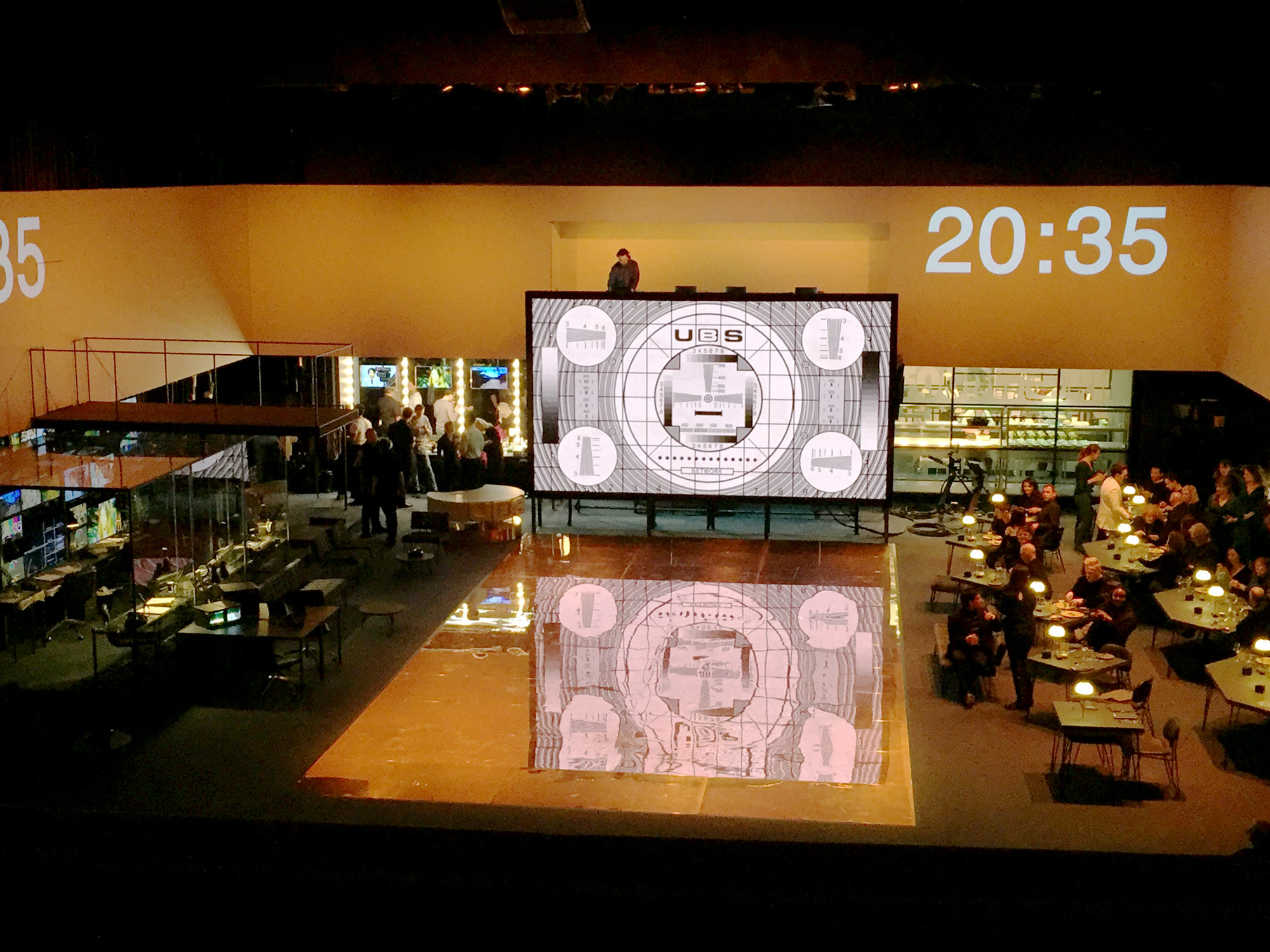 A large, open stage. To the right are several tables with people aorund them. To the rear a large screen shows a test-card, next to it a projected digital clock reads 20:35.