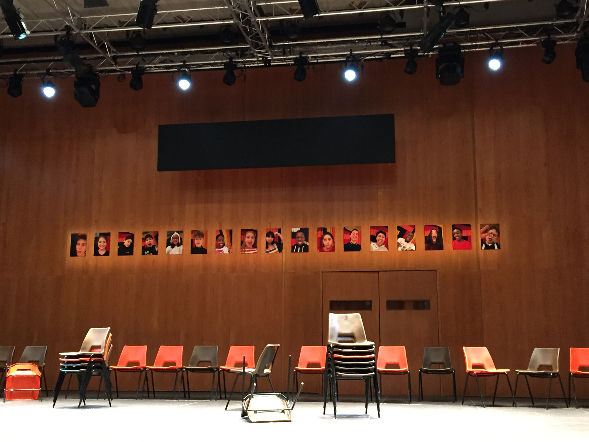 Dozens of plastic chairs are stacked and scattered on an otherwise empty stage. Behind them a wooden backdrop has 17 small photographs of children stuck in a row