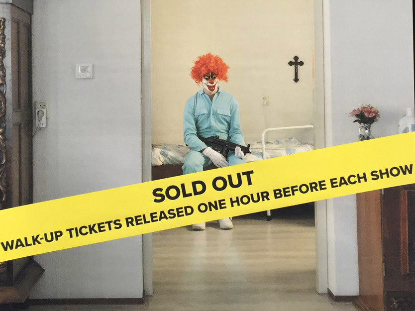 Through a doorway we glimpse a man dressed in a blue boiler-suit, a clown-mask and red wig. He is sitting on a bed holding a machine-gun. Across the whole image is a yellow strip reading Sold Out... 