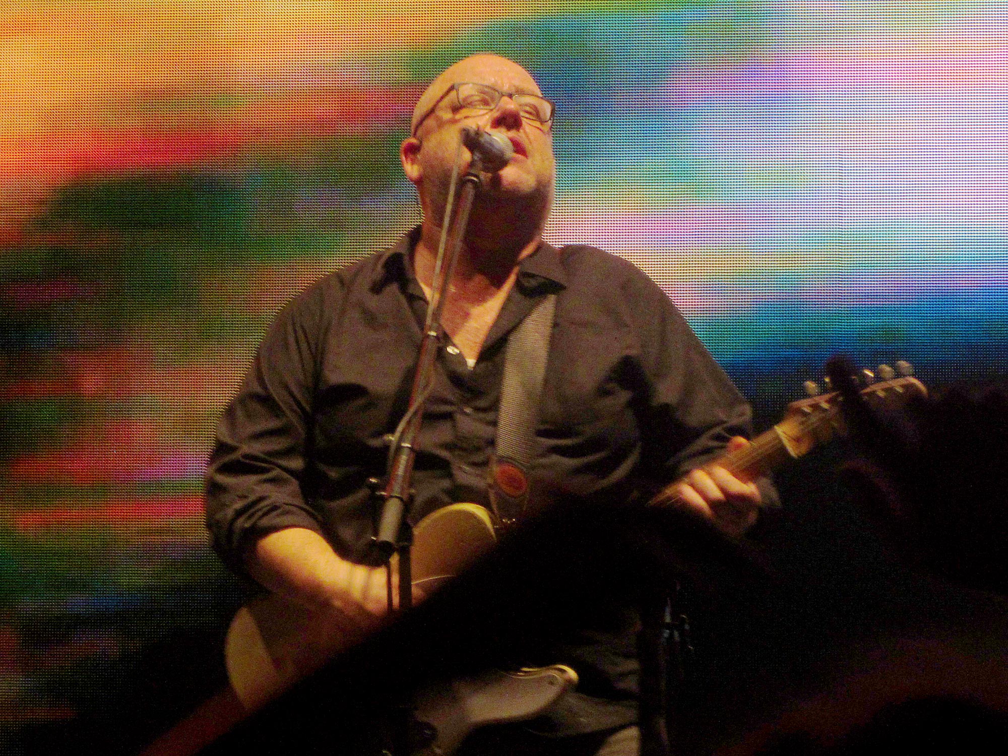 A bald man in glasses sings into a microphone whilst playing guitar. Behind him is a multicolour digital screen.