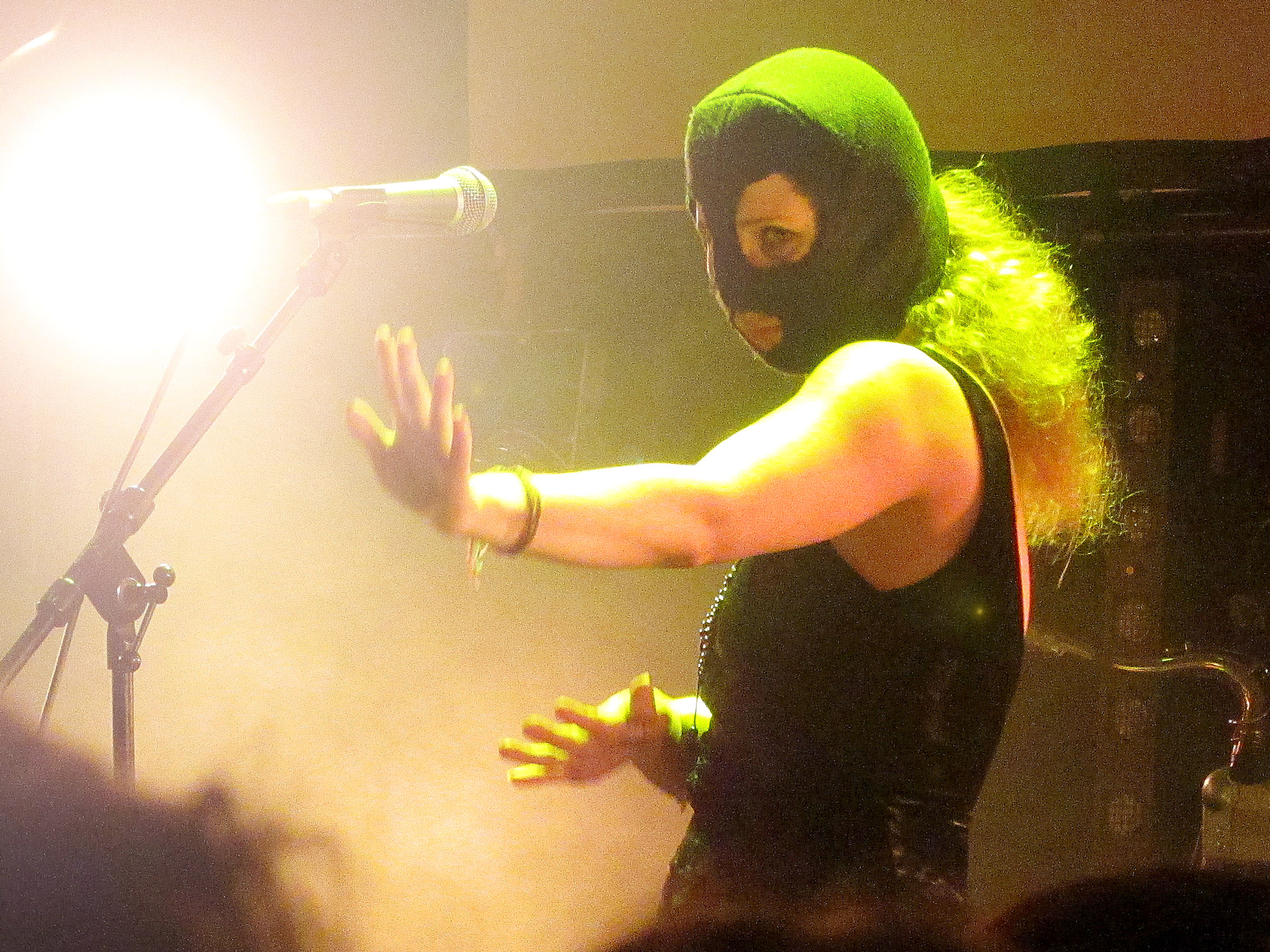A woman dressed in a sleeveless top is dancing on a misty stage, She is wearing a green balaclava, her hair billowing out of the back.