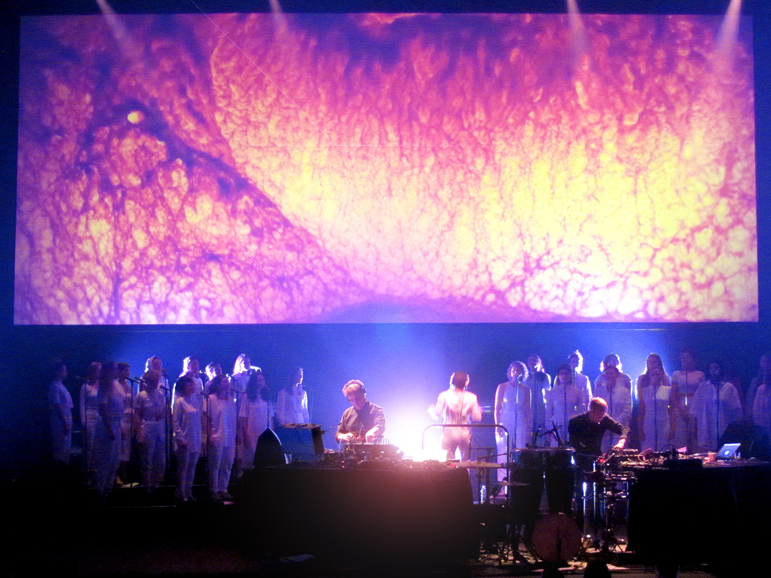Two DJs perform in front of an all-female choir. Behind them is an abstract image projection.