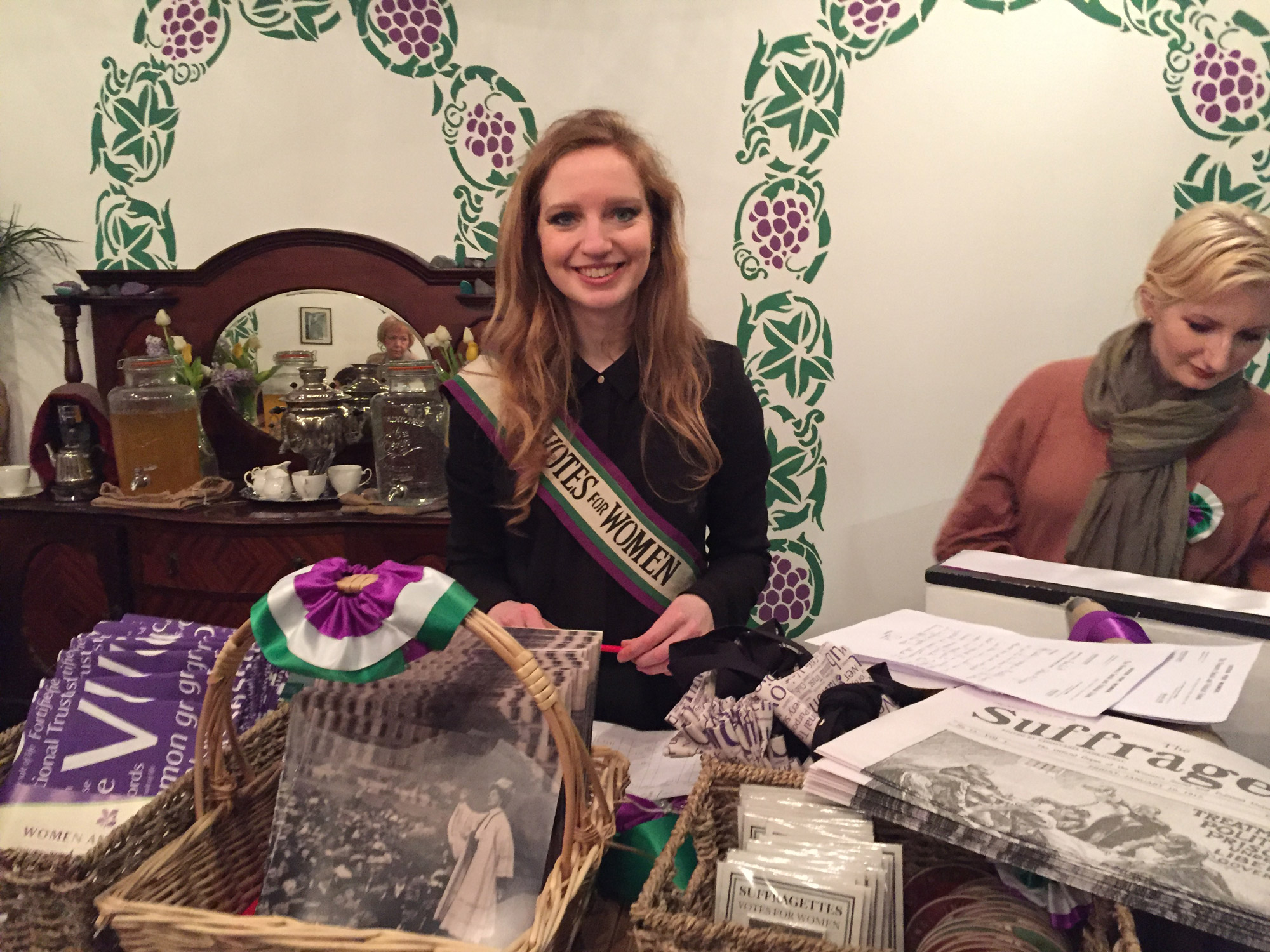 A woman, wearing a Votes for Women sash, smiles amongst items in a shop.