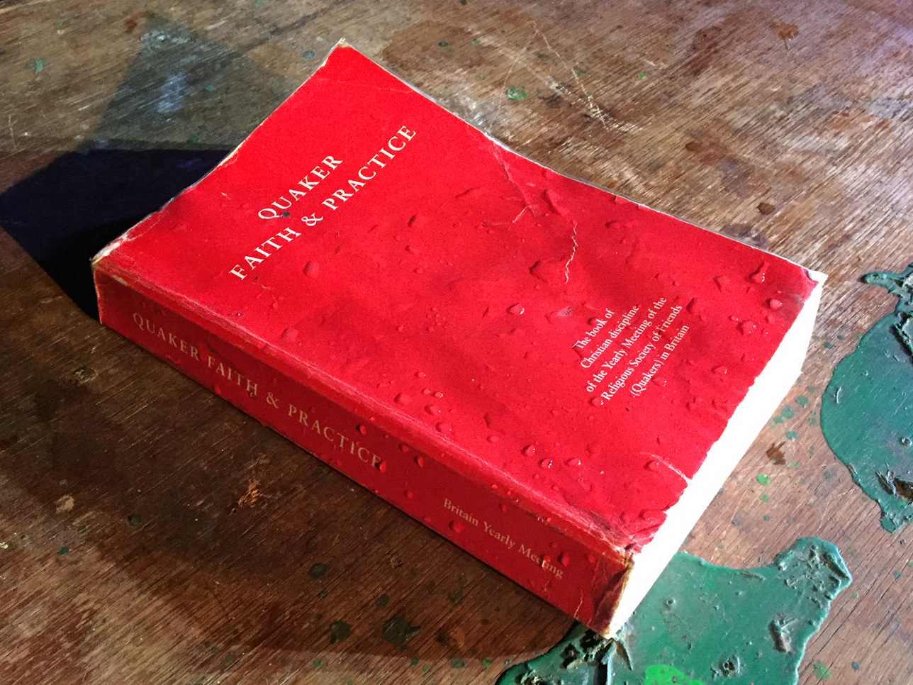 A water-soaked book, with a red cover, sits on a paint-splattered wooden surface.