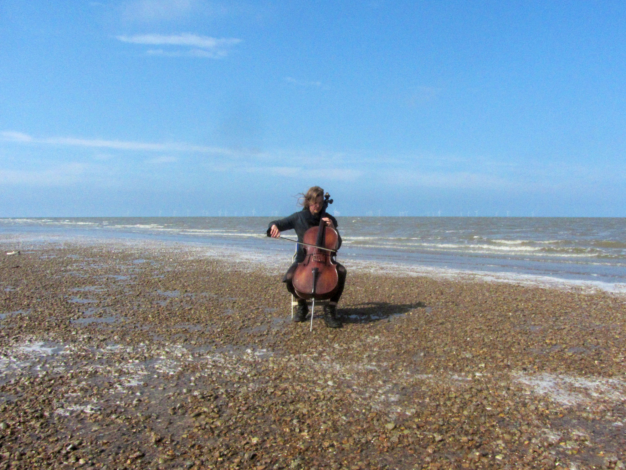 Under a blue sky, on a shingle beach, a woman plays a cello. Behind her we can see the sea.