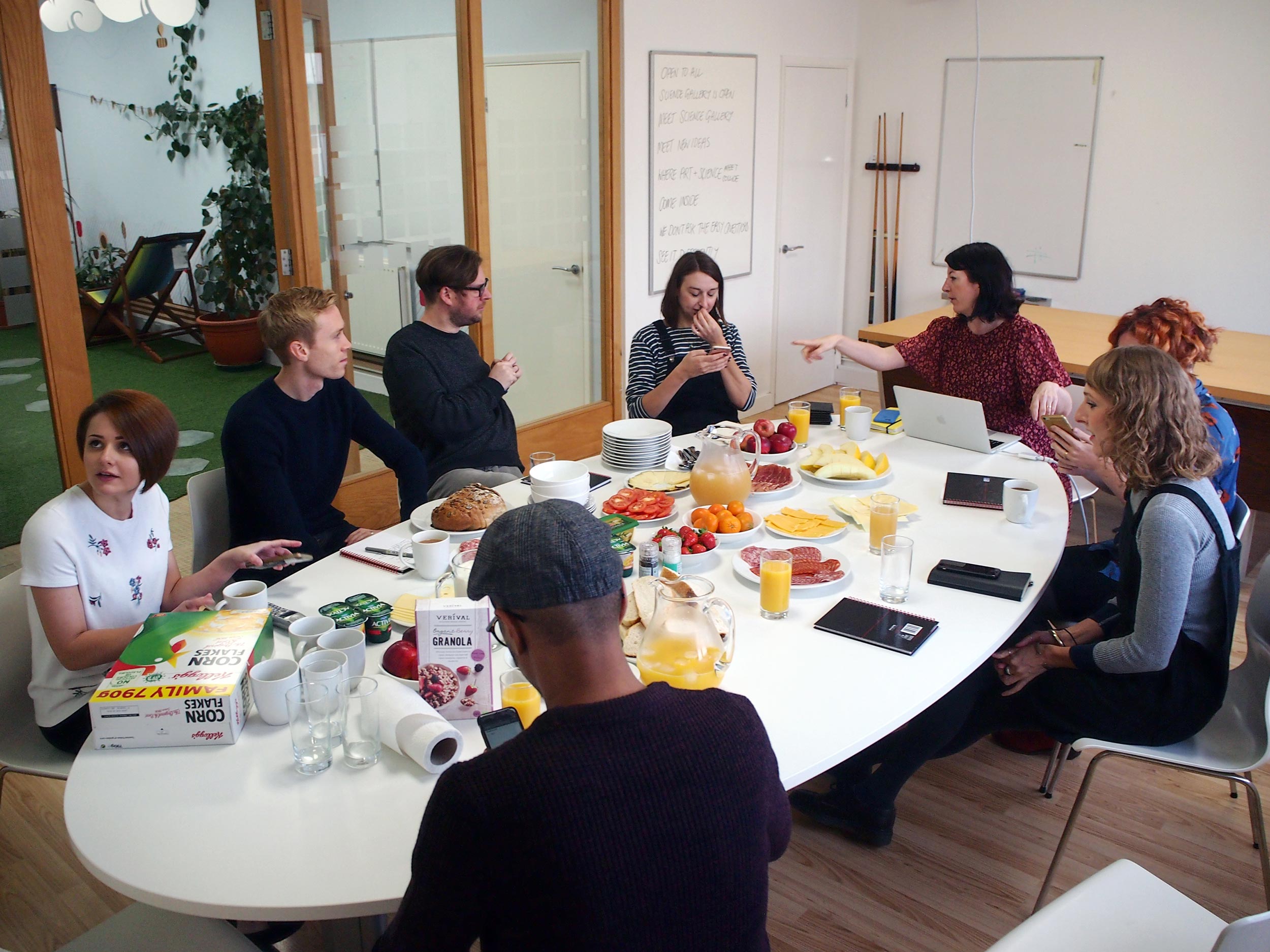 Eight people sitting around a white oval table, filled with fruit and other breakfast food