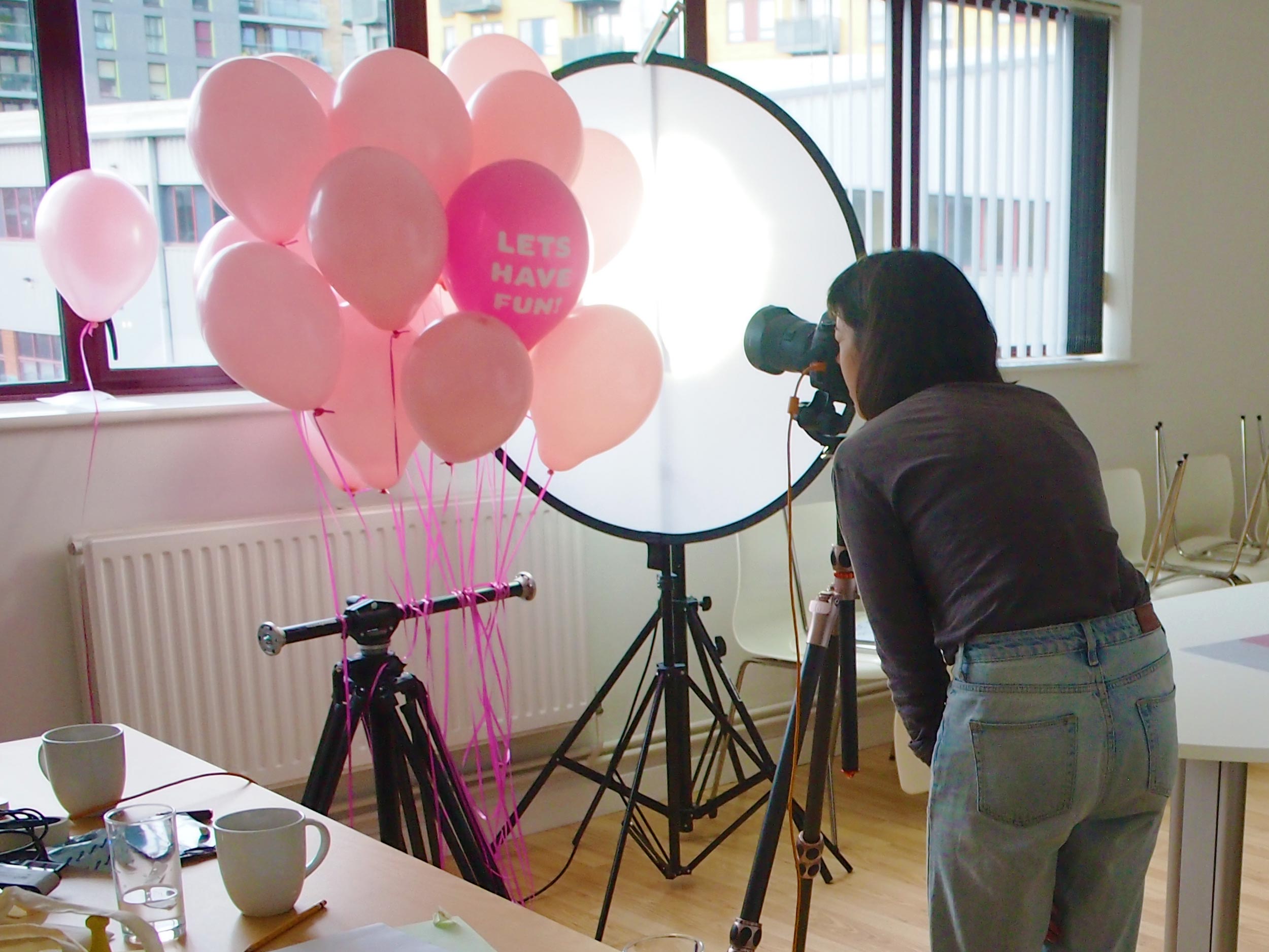 A photographer shoots pink balloons against the light of a window.