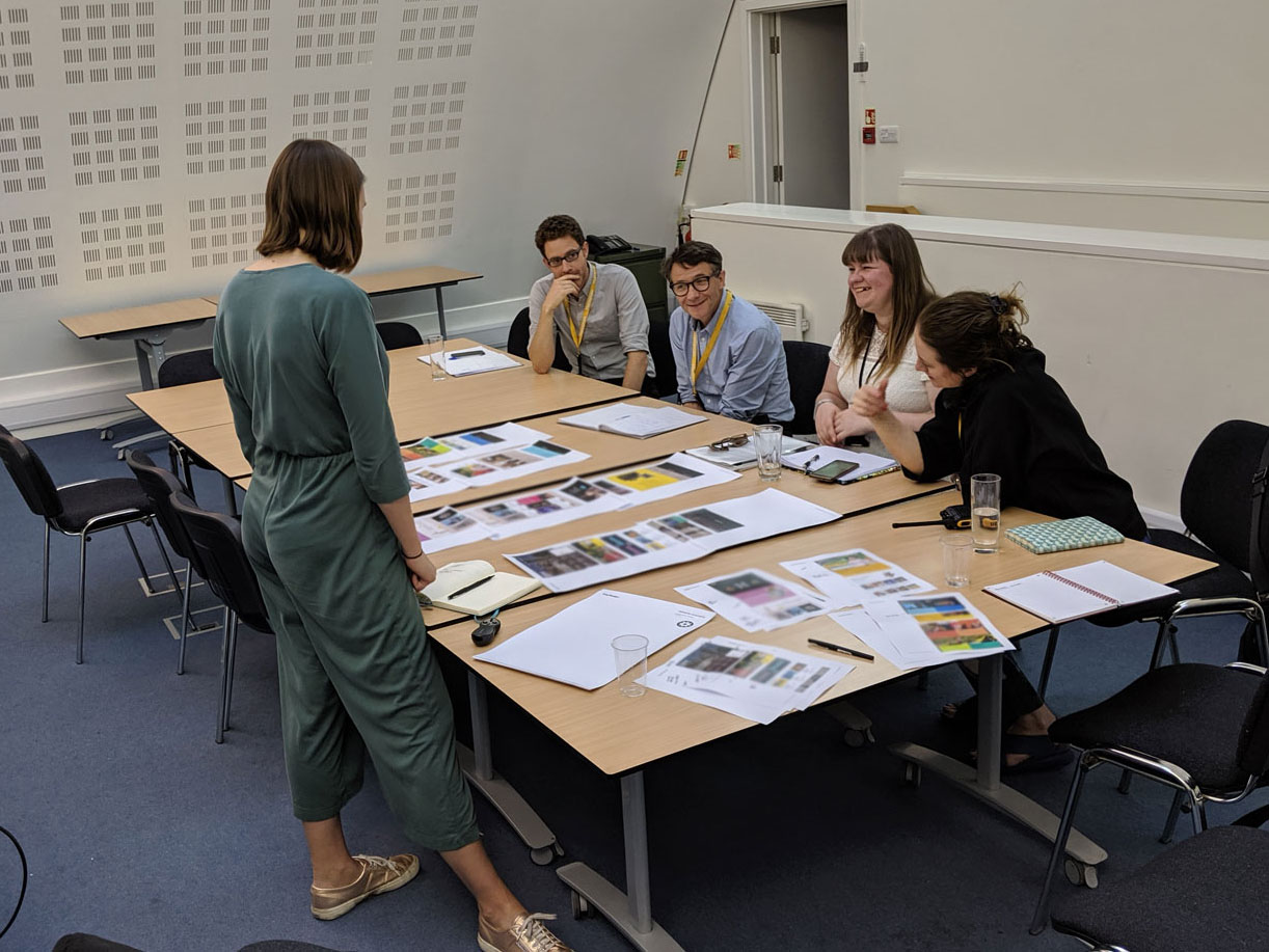 A woman stands over a table, four people look at pages, smiling