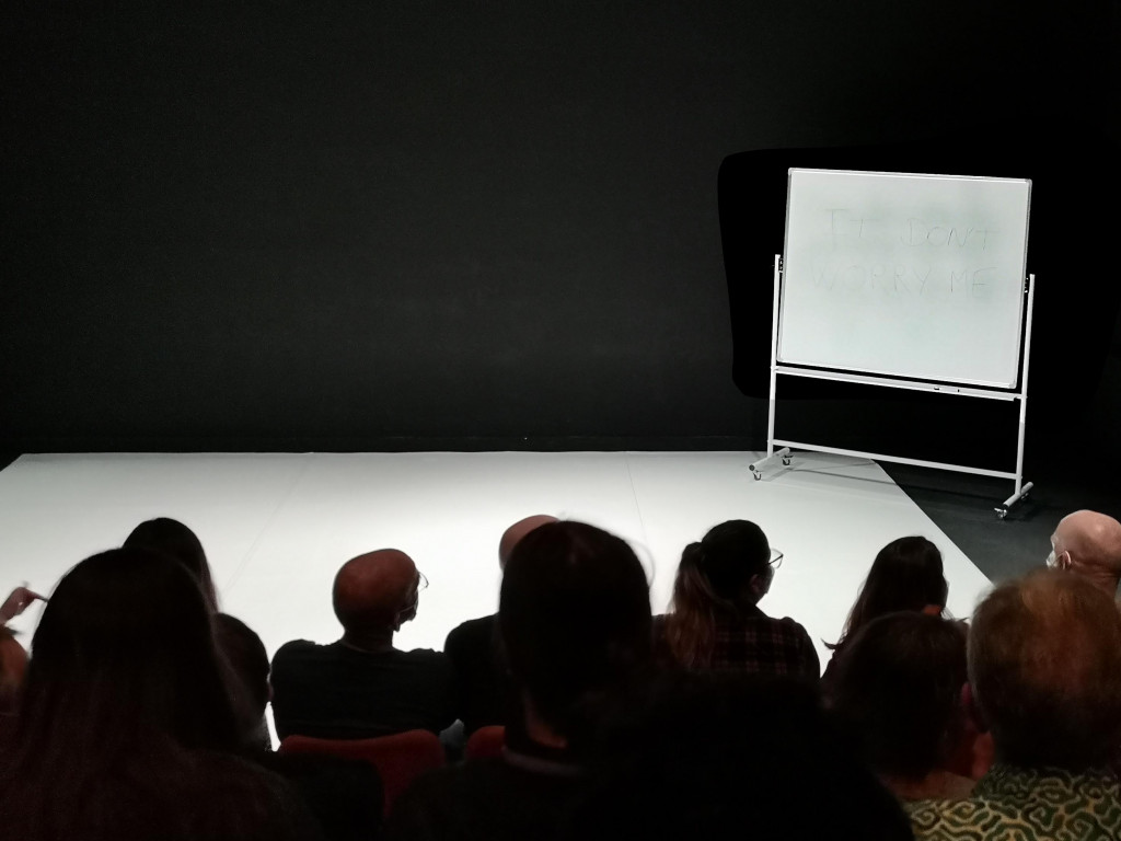 An audience look on to an empty performance space, in the corner is a portable white-board 