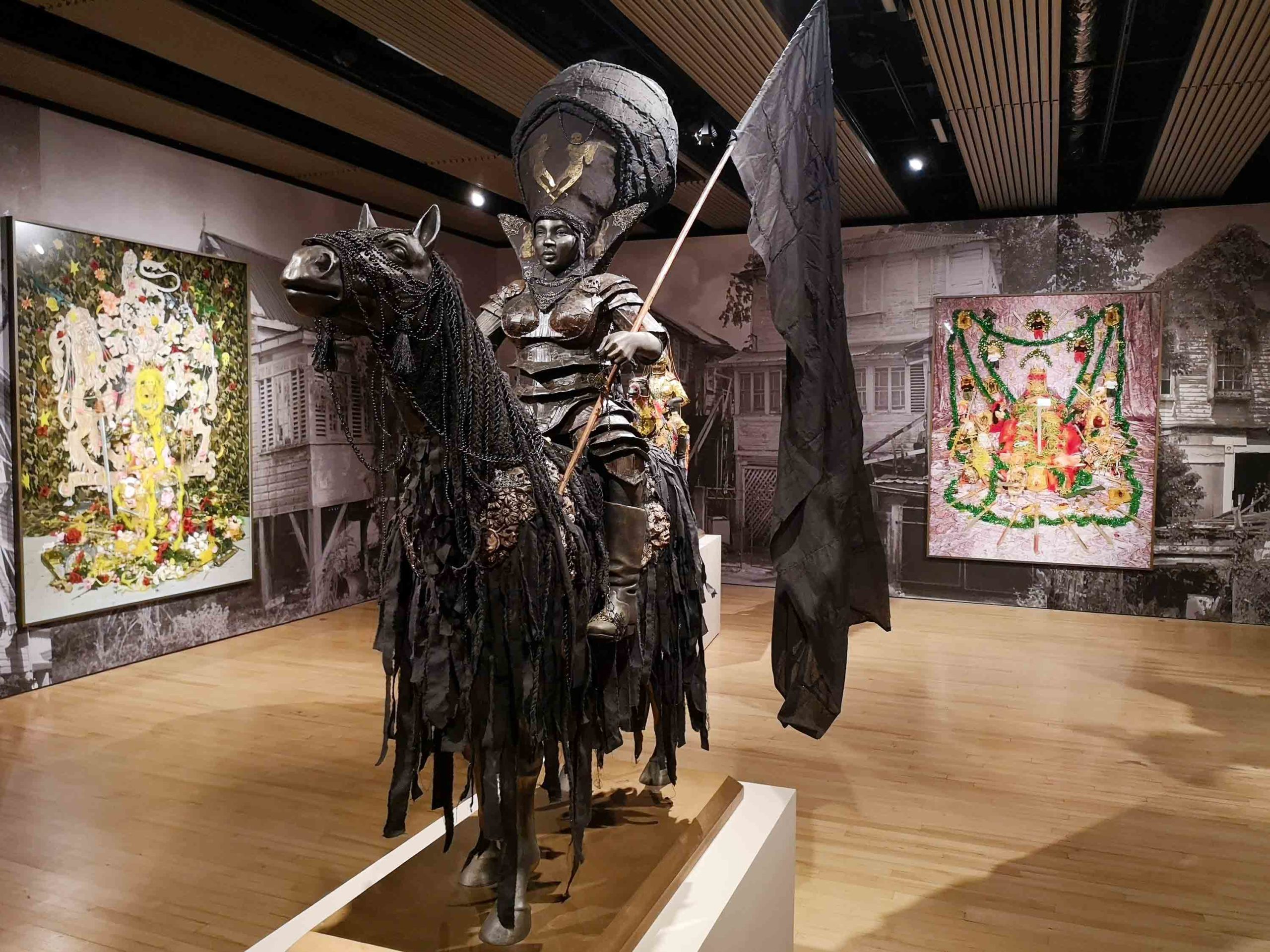 In a dark gallery with images on the walls. A plinth holds the statue of a black-clad figure on a black horse. The figure holds a black flag and is dressed in figure-hugging armour-like clothing, and a large bulbous head covering.