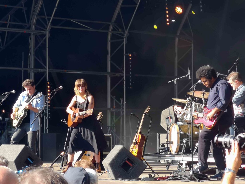 Daytime, we see four musicians playing on a stage. In the centre a woman with long brown hair wears a black dress and plays an acoustic guitar.