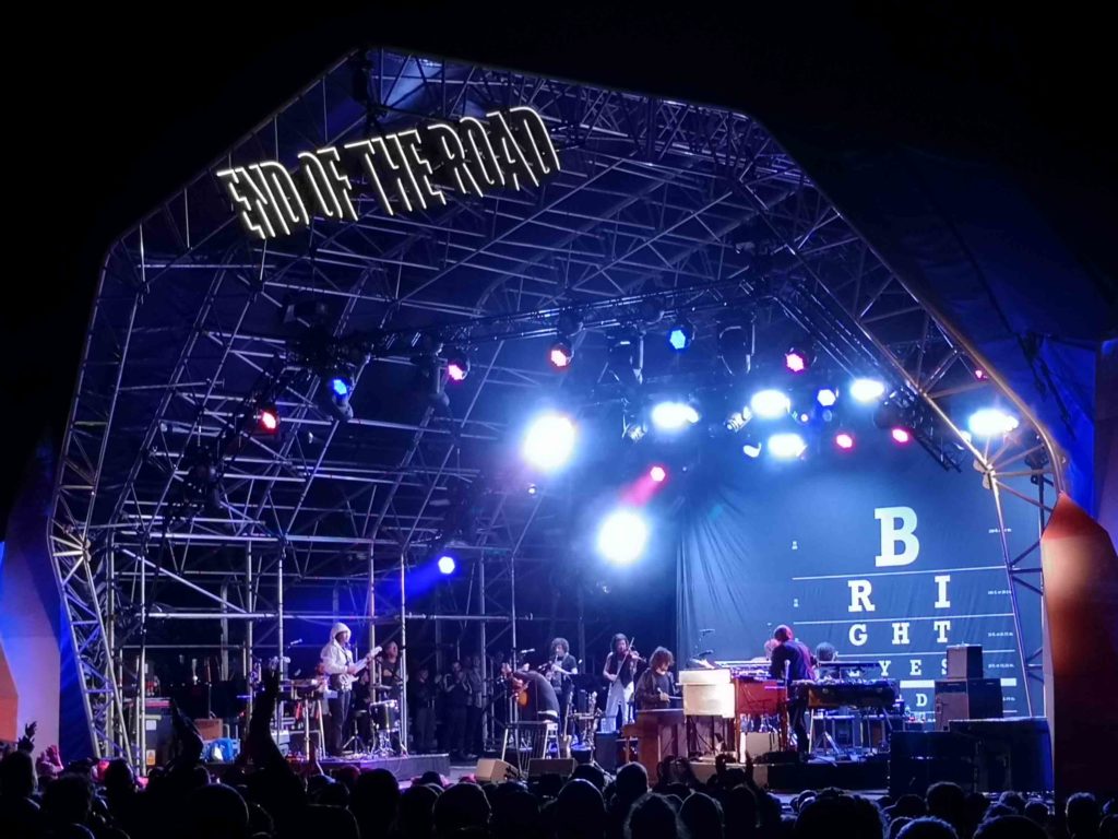 At night, a stage, covered in a dome-shaped cover is filled with musicians. In the foreground we see the heads of a crowd. Above the stage are the words END OF THE ROAD, at the back of the stage we can see the letters that spell out Bright Eyes, they are laid out on rows of descending sizes, like an eye-test.