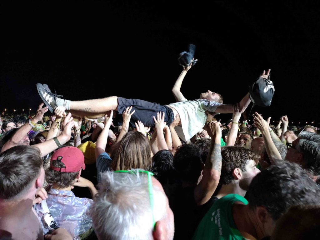 A young man, dressed in shorts and t-shirts lies on his back. He is held aloft by the multiple hands of the crowd.