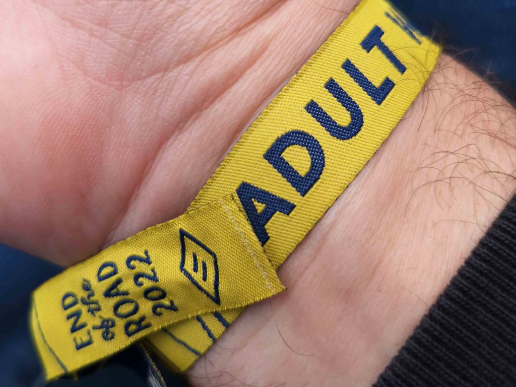 Close up crop of an in-situ wrist-band. The material band is yellow and features large blue type that reads ADULT. At the end of the band we can see the smaller type: END fo the ROAD 2022