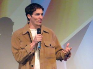 Chad Simpson. A casually dressed white man with shirt, dark hair, dressed in a light brown jacket. He speaks into a hand-held microphone.