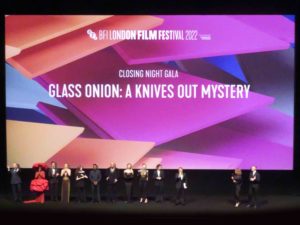 Glass Onion cast. Thirteen glamorously dress people stand in a line on a large stage Above them a projection reads: Glass Onion a Knives Out Mystery.