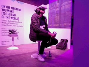 Nick Kime. A young white man sits of a white stool. He is wearing a coat over a dark jumper, dark trousers and white trainers. On his head he is wearing a Virtual Reality headset and headphones and is holding a controller. 