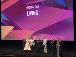 Eight smartly dressed people stand on a stage. In the centre we see a short man in a dark suitm speaking into a microphone while other look toward him. Behind them a projection reads Heading Gala Living.