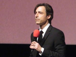 Noah Baumbach, a middle aged white man with straight brown hair. He is holding a microphone and is dressed in a black dress jacket, white shirt and black tie.
