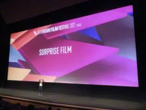 Michael Blyth. A casually dressed, lone young white man with shaved head stand, with a microphone in the middle of a large stage. Behind him is projected the words Surprise Film.
