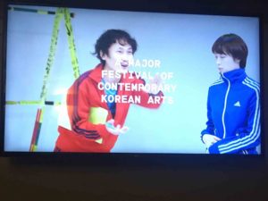 Screen projection shows two Korean men, dressed in tracksuit tops – one red, one blue. Overlaid on the image is type that read Major festival of contemporary Korean arts
