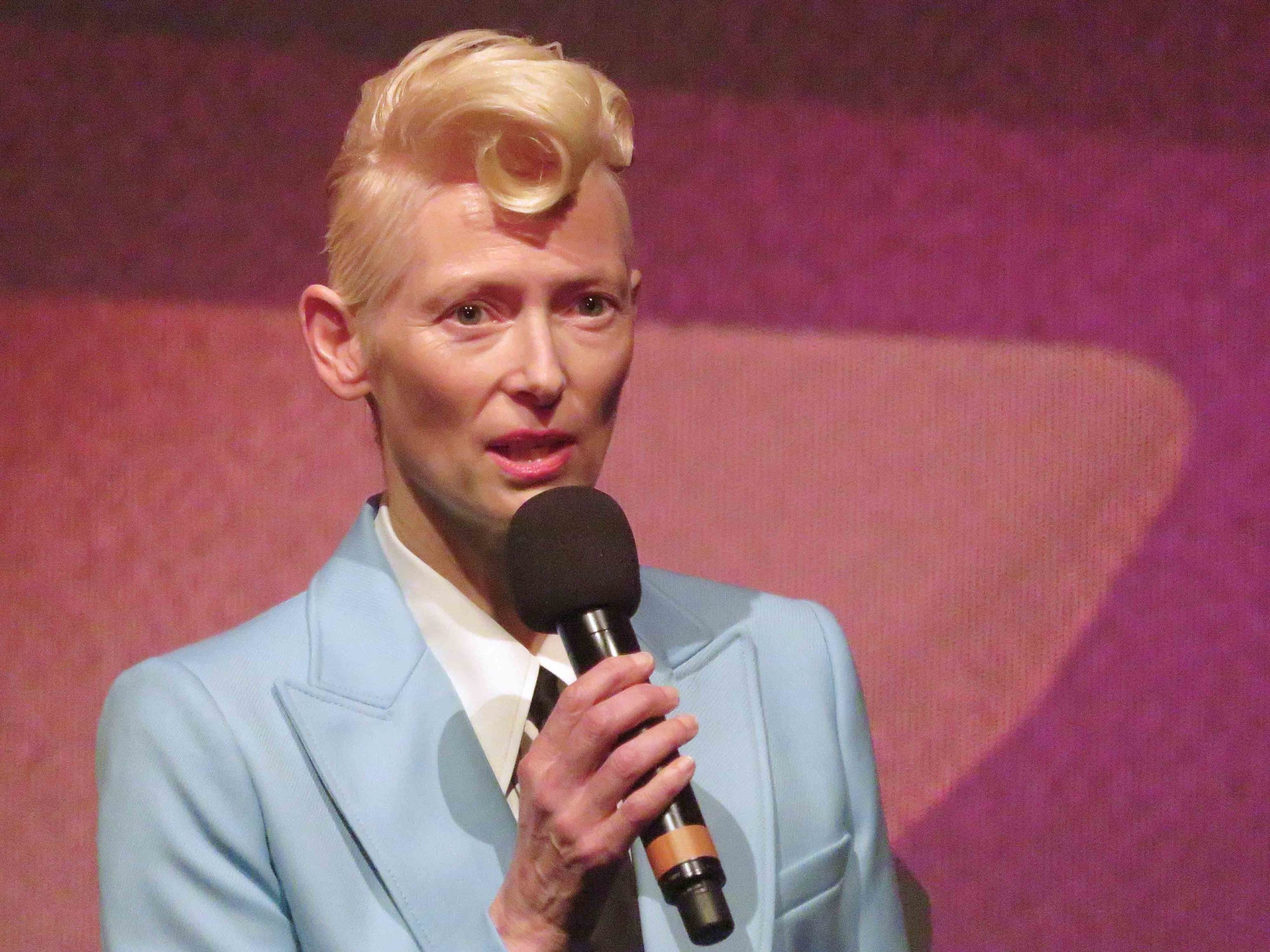 Tilda Swinton. A middle aged white woman. She has blond hair, shaved at the side and longer on top with a kiss curl fringe. She is holding a microphone and wearing a powder blue suit jacket, white shirt and black tie.