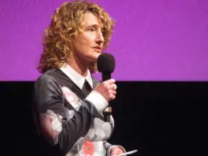 Tricia Tuttle. A middle aged white woman with long hair in curls. She is speaking into a microphone and is wearing a dark dress with a flower pattern and white cuffs and collar.