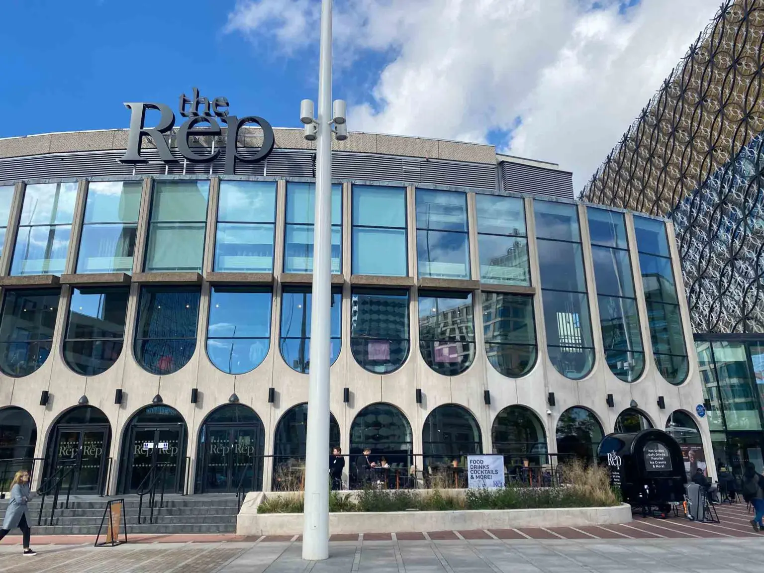 Against a blue sky, a large modernist building with large windows, in a pedestrian area. Atop the building a sign reads 'the Rep'