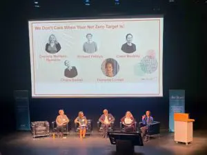 From a distance, we see five seated people in a row, on a stage. Above and behind them, a screen shows their names and photos in circles.