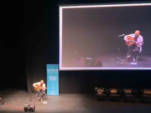 A bald man, with a beard, sits on a large stage, playing a stringed instrument (an oud). Behind him is a large live projection of him.