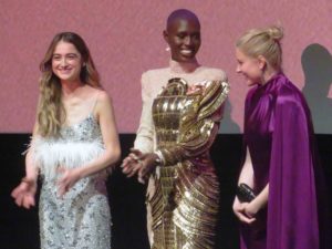 Raffey Cassidy, Jodie Turner-Smith and Greta Gerwig. Three glamorously dressed young women stand in a line, laughing. 