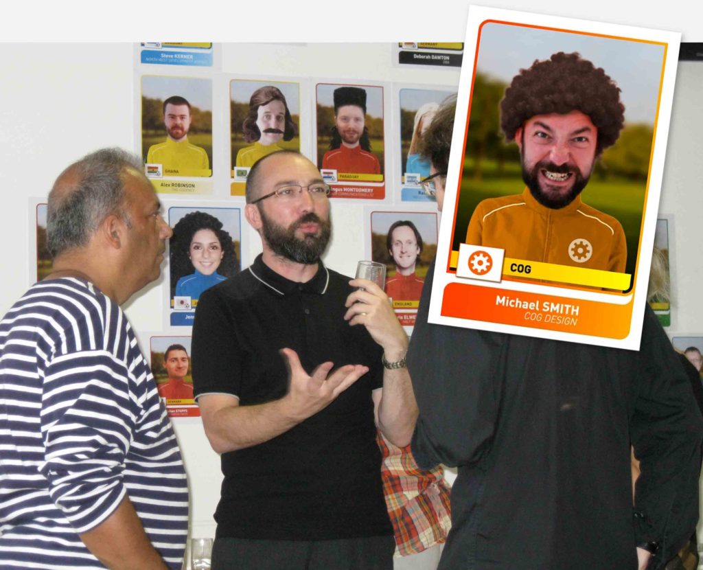 Three middle aged men are standing, talking. On the wall behind are print outs of giant pastiche football stickers. Overlaid on the image is anotehr pastiche football sticker, this one features Michael Smith in an orange tracksuit top. A perm-style wig has been added on his head. 