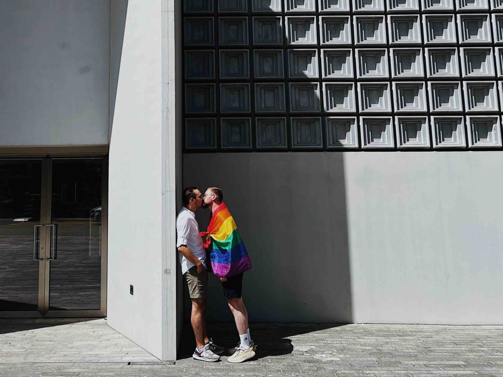 In front of a modern, concrete building, two young men kiss. Both are wearing shorts, one is draped in a rainbow flag.