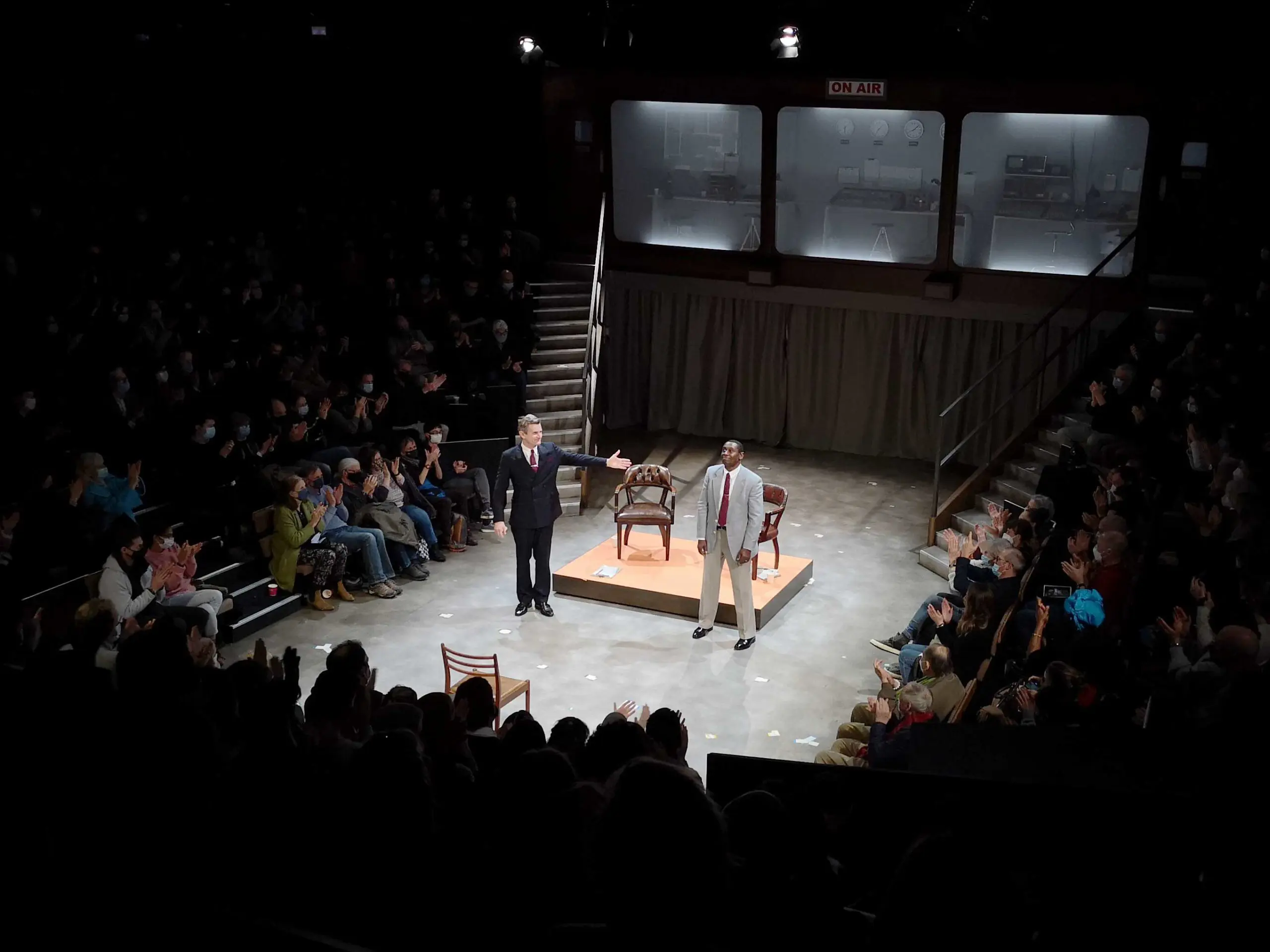 Charles Edwards a white man, and David Harewood a black man, stand on a stage surrounded on three sides by banks of seated audience. The men are dressed in suit and tie, their is a tiny raised stage with two chairs behind them.