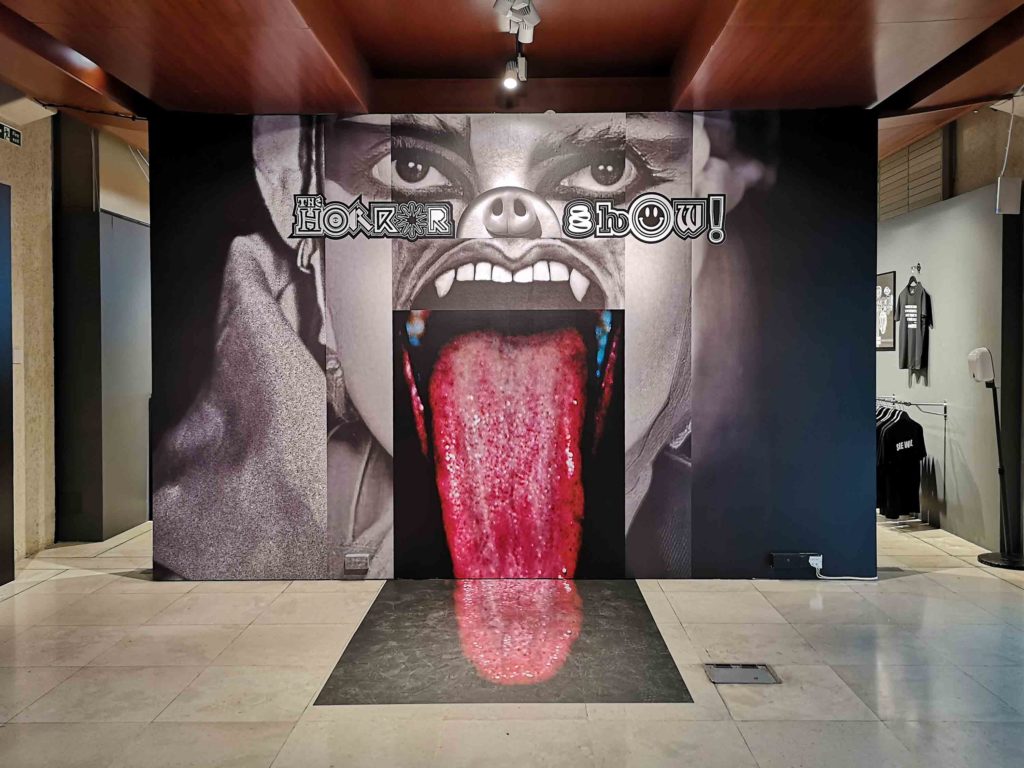 Printed floor to ceiling display shows a grotesque face, collaged from different images. At the centre, an open mouth, with fangs and a tongue that extends out of the picture and across the floor.