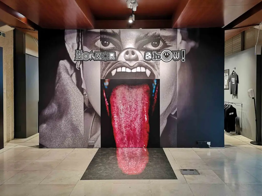 Printed floor to ceiling display shows a grotesque face, collaged from different images. At the centre, an open mouth, with fangs and a tongue that extends out of the picture and across the floor.