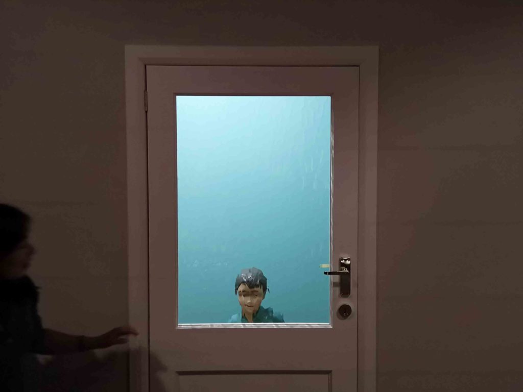 Behind the frosted glass of a closed door we see the distorted image of a child. In front, a shadowy figure is reaching to open the door.