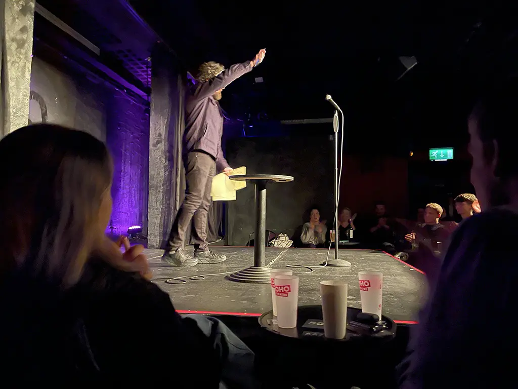 Alfie Brown stands on the stage, waving goodbye to the crowd. The audience are sat down looking up at him applauding.