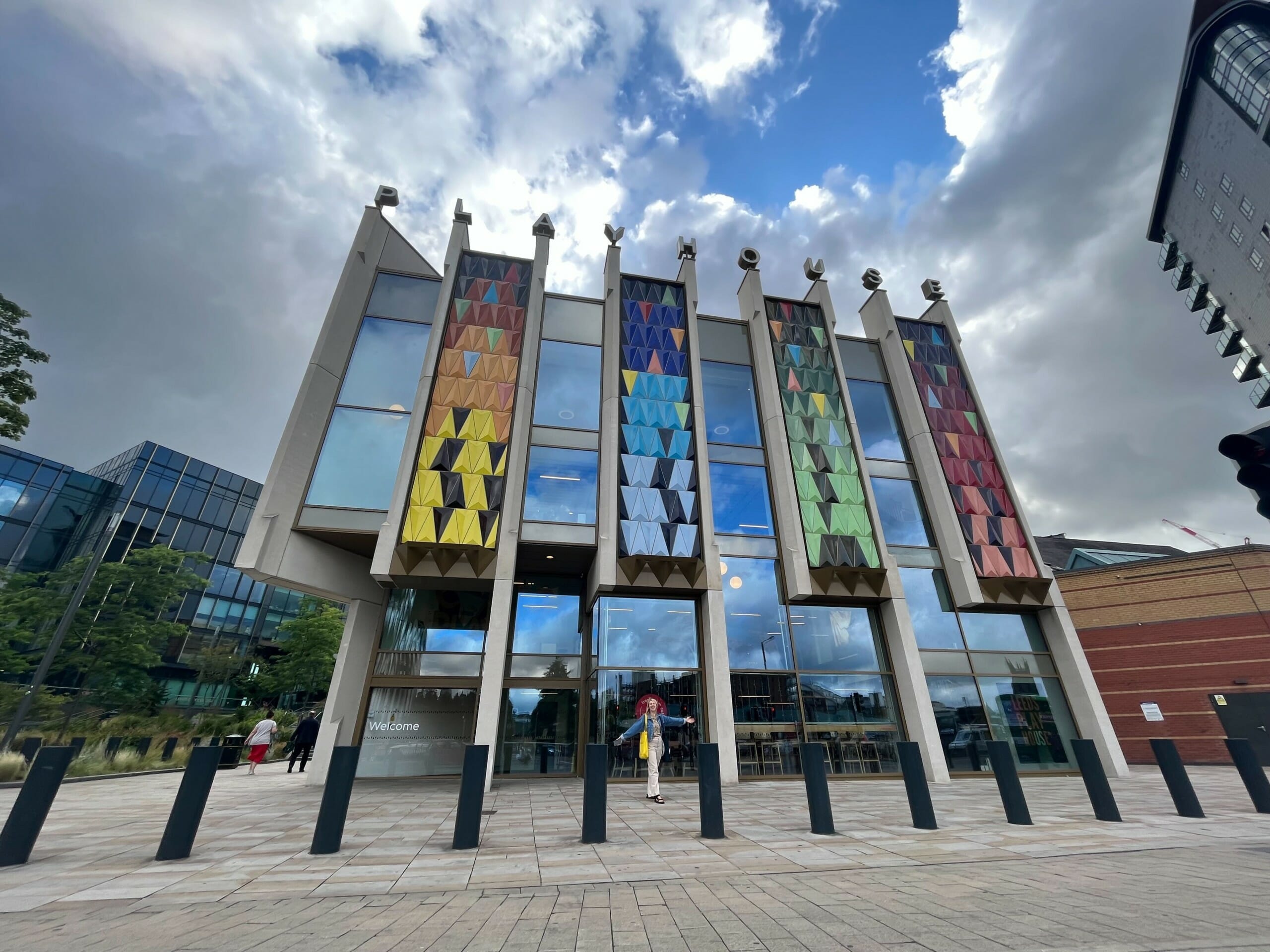 Emma, a blonde haired woman wearing a denim jacket, stands with her arms out wide in front of the Leeds Playhouse. The bright coloured facade of the building and sunshine is reflected onto the glass windows of the building.