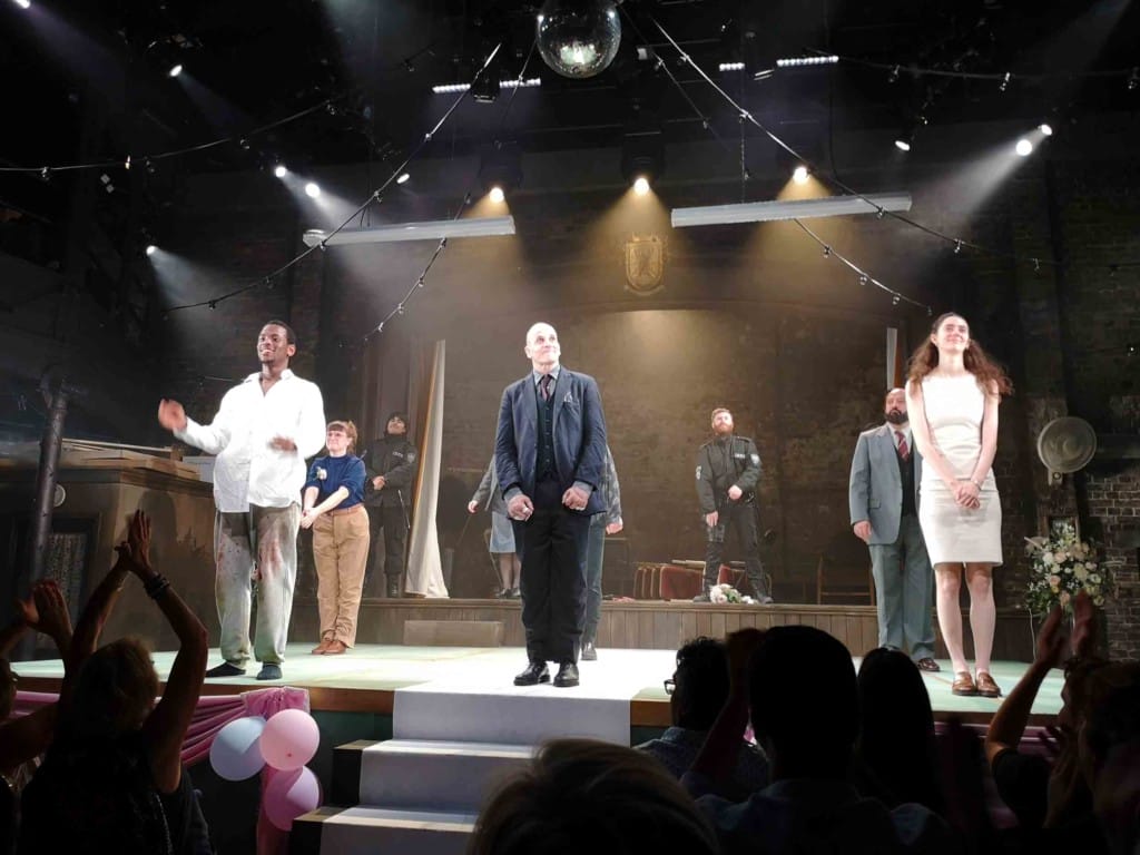 Eight actors stand on a stage in rows. The stage has steps at the front, flanked by balloons.