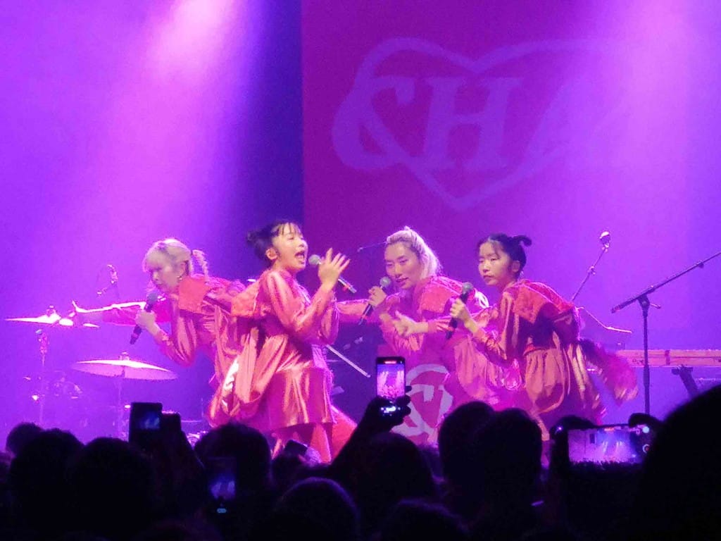 Four Japanese women singers, dressed in pink silky costumes hold poses whilst singing, side on to the front of the stage. 
