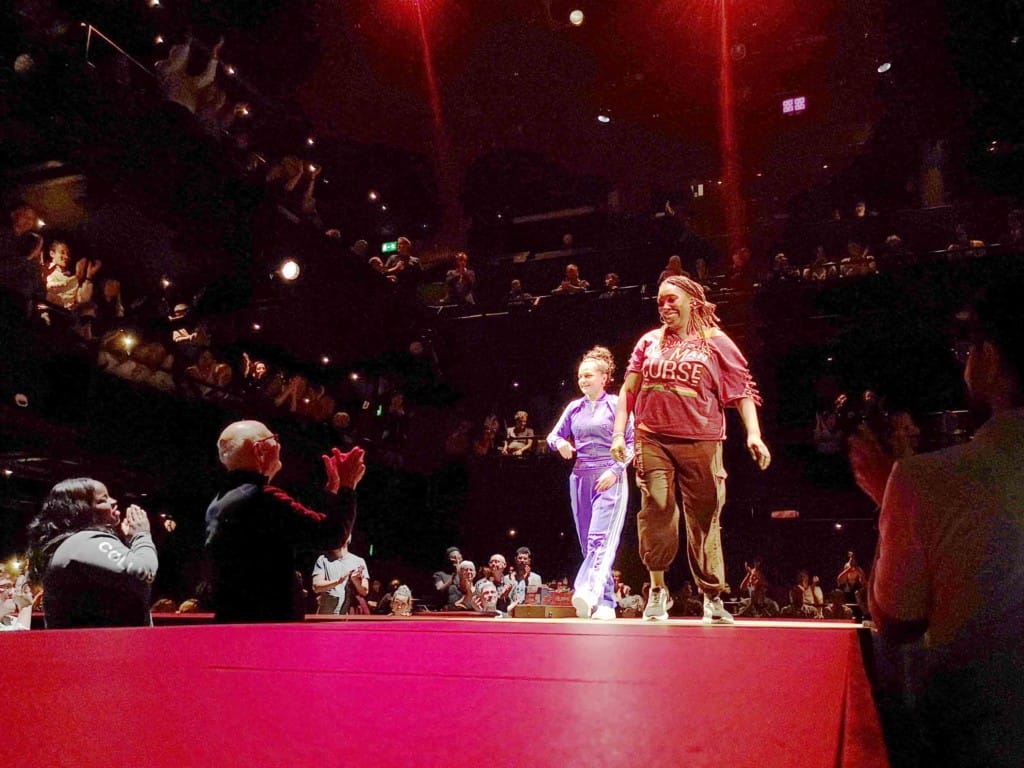 From a low angle, looking up from a red surface we see two casually dressed female actors, one white, one brown skinned, taking applause from a crowd in staggered seating. 