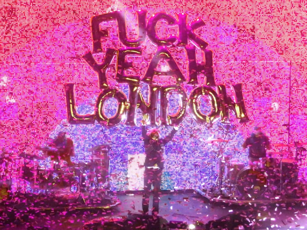 A foreground filled with confetti and the background filled with an LED screen with a purple and pink rainbow shape. Through the confusion we see two drummers behind drum kits, and in the centre, a man holds his arms aloft. He is holding gold balloons that spell out: fuck yeah London.